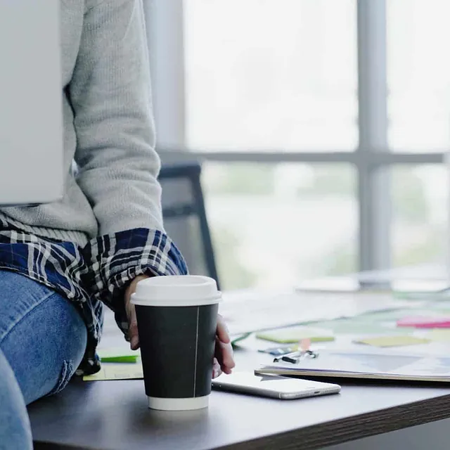 Person i jeans og rutete skjorte holder en kaffekopp, sittende på et skrivebord med laptop